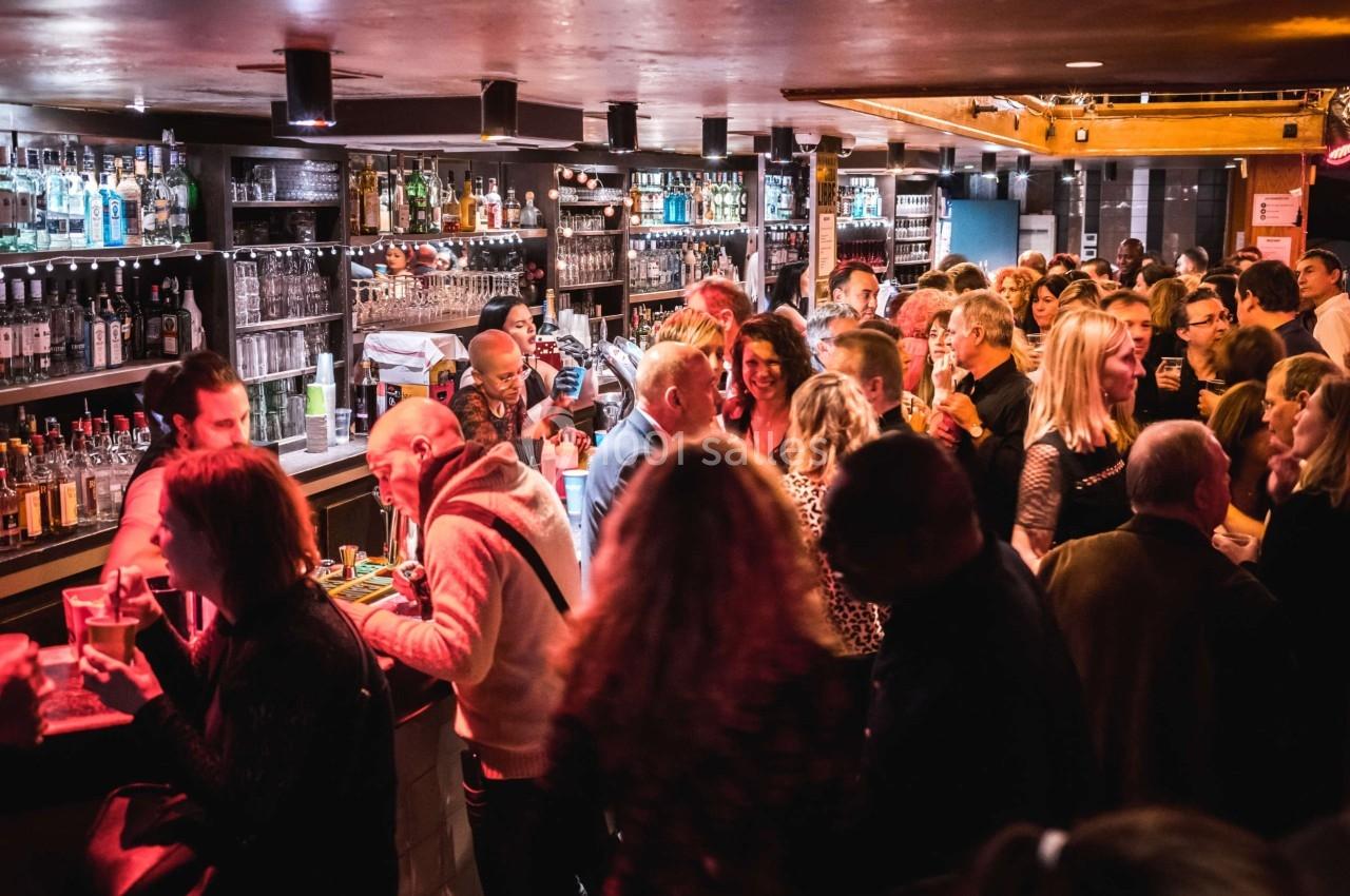Groupe de personnes discutant et buvant dans un bar animé avec un éclairage tamisé.