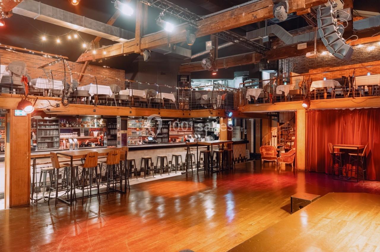 Salle avec bar central, tables en mezzanine, éclairage chaleureux et scène en bois dans un décor industriel.