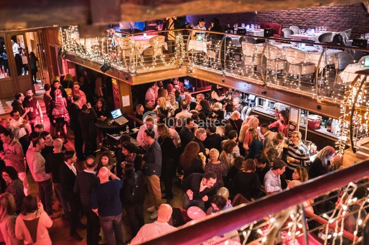 Salle bondée avec des personnes debout et discutant, éclairée par des guirlandes lumineuses et un éclairage chaleureux.