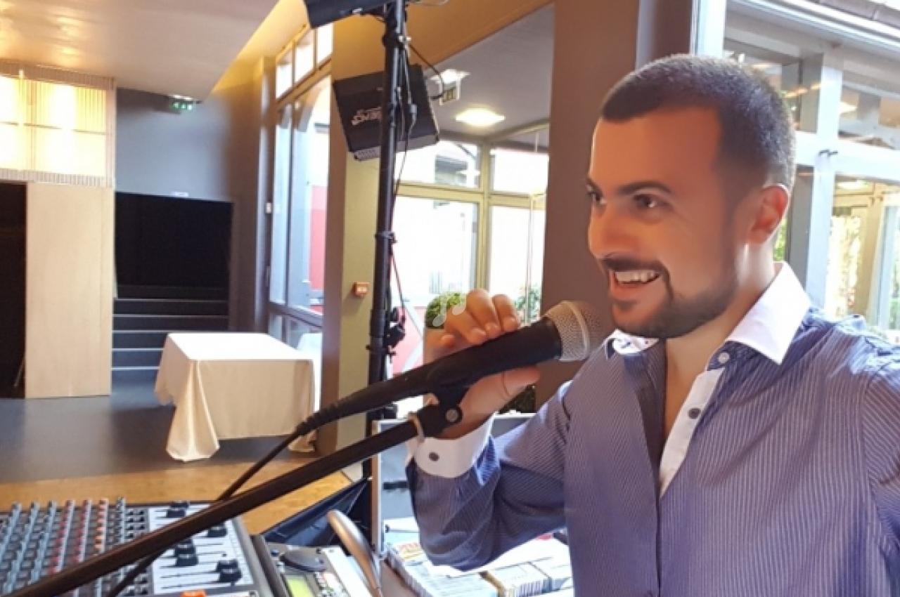Un homme souriant parle dans un micro devant une table de mixage dans un espace intérieur lumineux.
