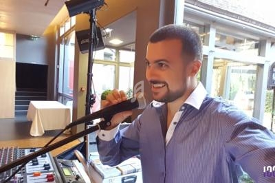 Homme souriant parlant dans un micro, devant une table de mixage, dans une salle lumineuse avec de grandes fenêtres.