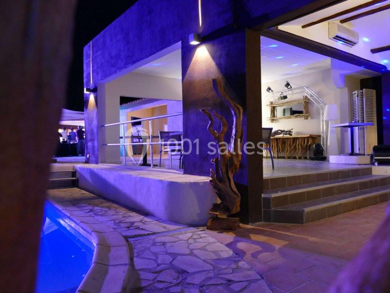 Terrasse moderne éclairée de nuit avec piscine, mobilier extérieur et sculpture en bois près d'une entrée ouverte.