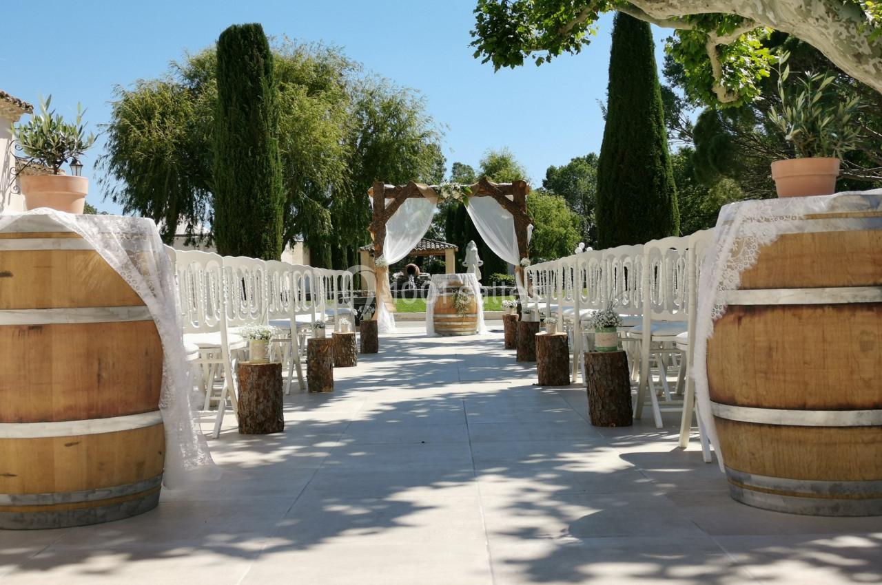 Allée extérieure décorée pour une cérémonie de mariage, avec chaises blanches, tonneaux en bois et arche ornée de voilages.
