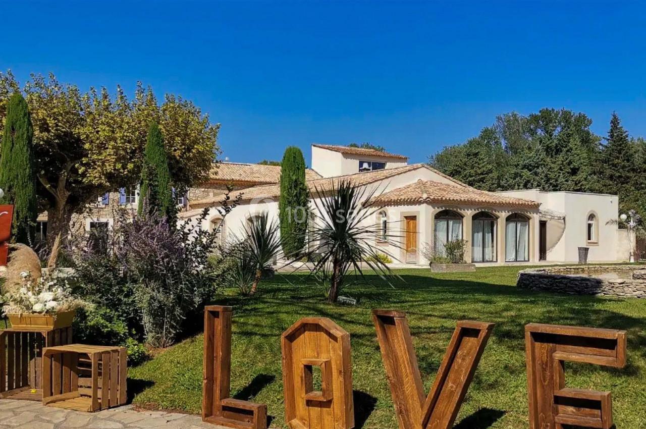 Jardin avec pelouse, arbustes et sculptures en bois formant le mot ’LOVE’, devant une grande maison méditerranéenne.