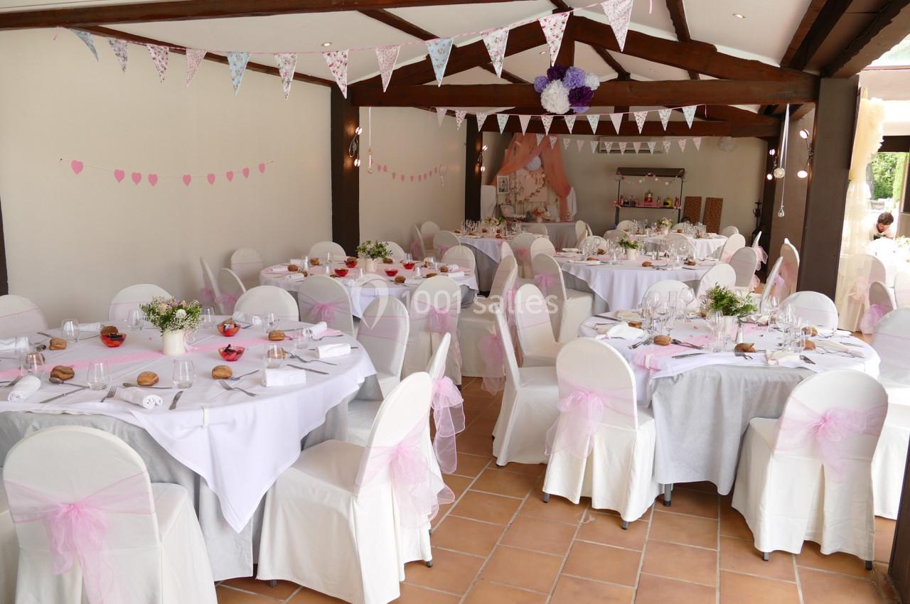 Salle décorée pour une réception avec des tables rondes dressées, des chaises blanches ornées de nœuds roses et des…