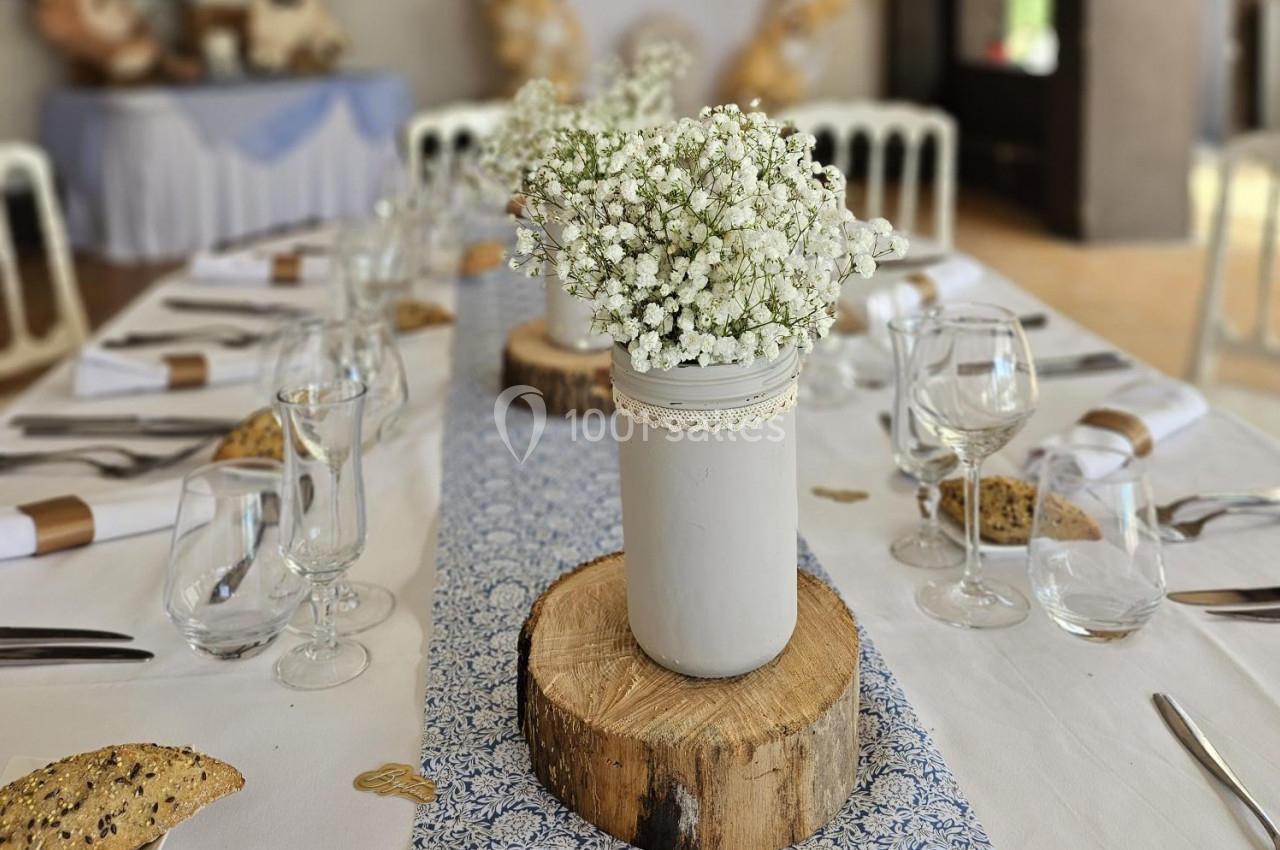 Centre de table avec un vase blanc rempli de gypsophiles, posé sur une rondelle de bois, entouré de couverts dressés.