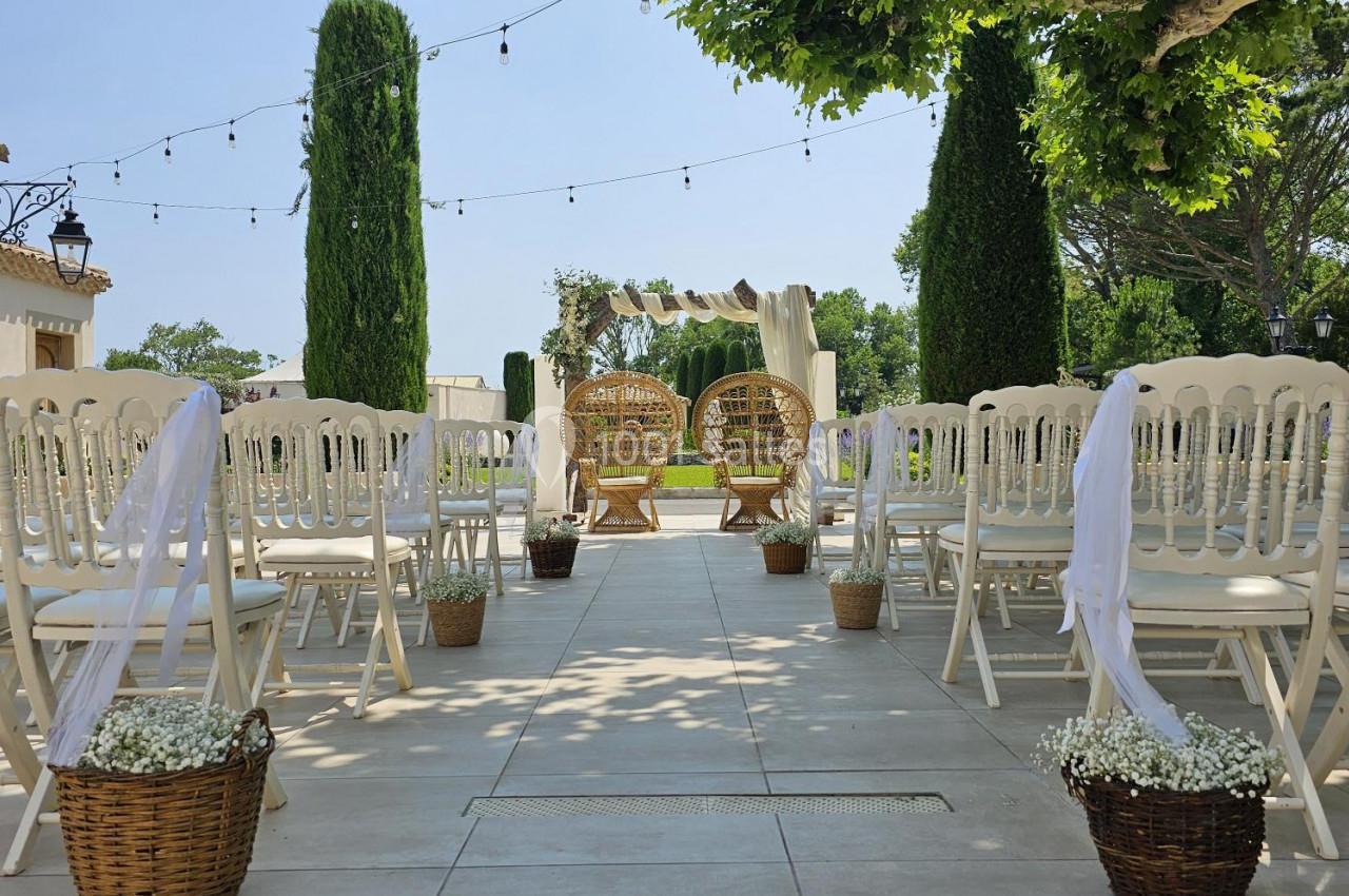 Allée centrale décorée pour une cérémonie en extérieur, entourée de chaises blanches et de paniers fleuris.