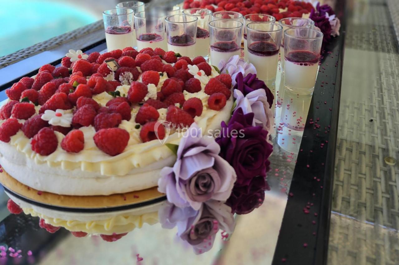 Gâteau décoré de framboises et fleurs violettes, accompagné de verrines de dessert sur une table en verre.