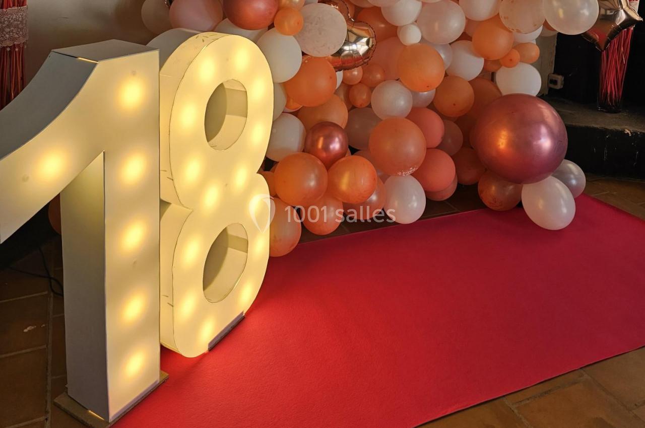 Chiffre 18 lumineux devant un mur de ballons roses, blancs et orange, avec un tapis rouge au sol.