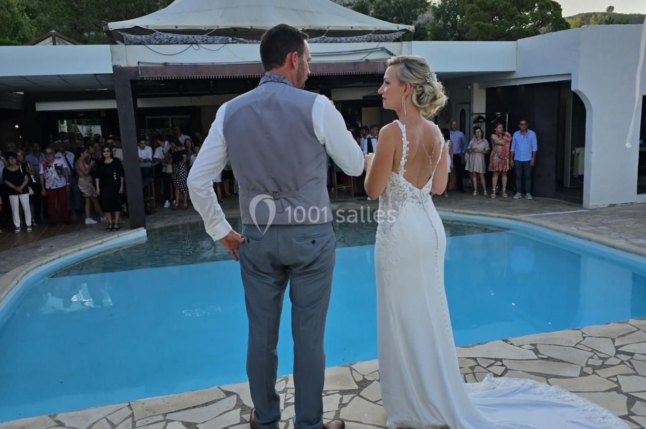 Un couple de mariés debout près d'une piscine, face à des invités rassemblés sous une pergola.