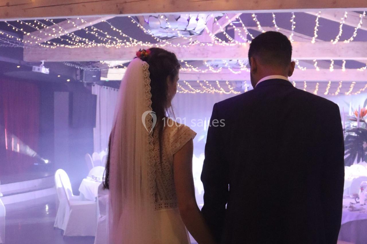 Un couple de mariés vu de dos dans une salle décorée de guirlandes lumineuses et de tables dressées.