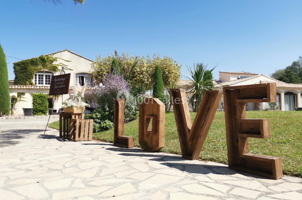Lettres en bois formant le mot ’LOVE’ disposées sur une allée pavée devant une maison avec jardin.