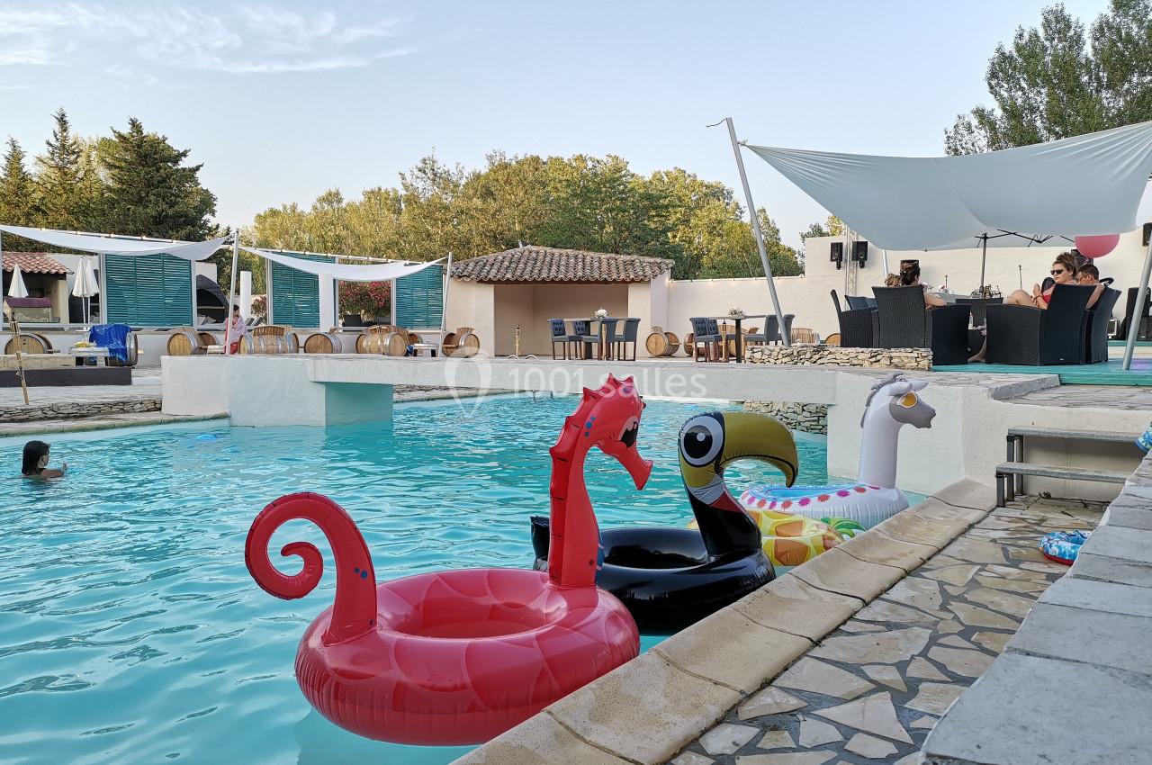 Des bouées colorées en forme d'animaux flottent dans une piscine extérieure entourée de terrasses et de végétation.