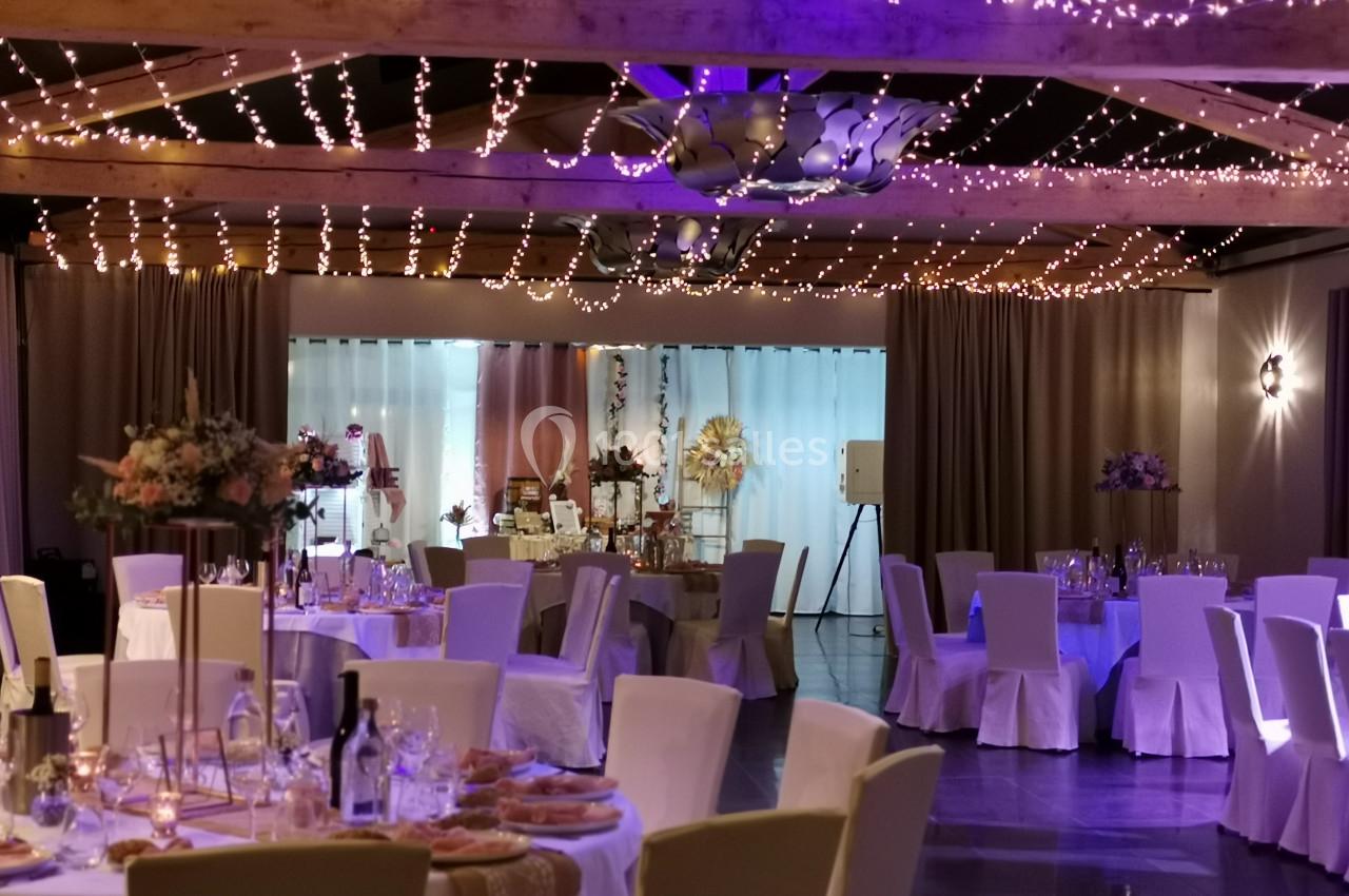 Salle de réception décorée avec des guirlandes lumineuses, tables dressées avec nappes blanches et compositions florales.