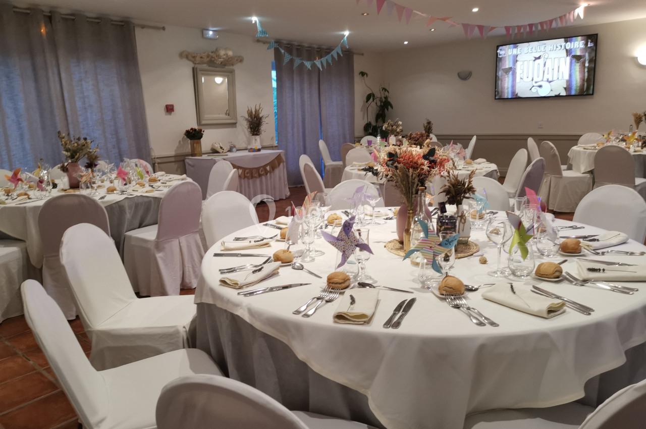 Salle de réception décorée pour un événement, avec tables rondes dressées, chaises blanches et décorations florales.