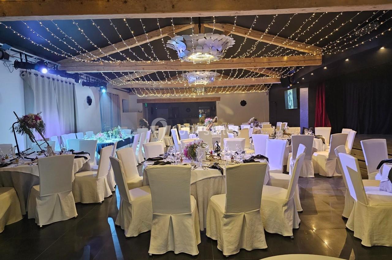 Salle de réception décorée avec des tables rondes dressées, des chaises blanches et un plafond illuminé de guirlandes…