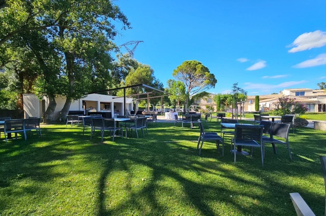 Espace extérieur avec tables et chaises sur une pelouse, entouré d'arbres et de bâtiments sous un ciel bleu.