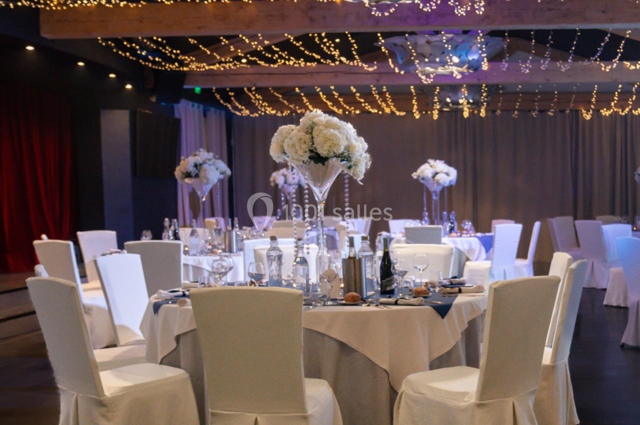 Salle de réception décorée pour un mariage avec tables rondes, nappes blanches, centres de table fleuris et guirlandes…