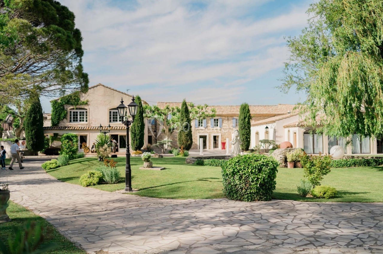 Bâtisse en pierre entourée d'un jardin verdoyant avec allée pavée, arbres et arbustes sous un ciel dégagé.