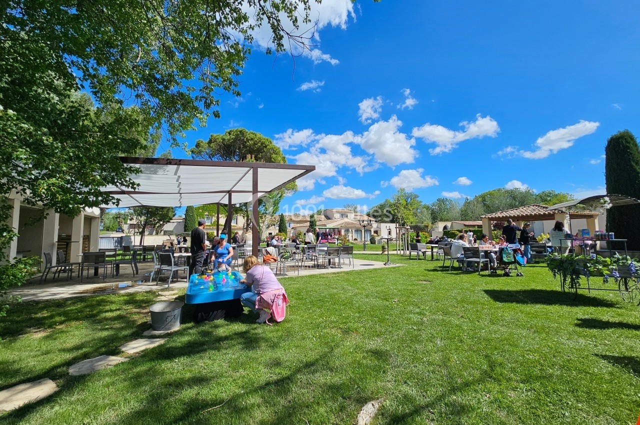Personnes profitant d'un espace extérieur verdoyant avec tables, chaises et activités sous un ciel bleu.