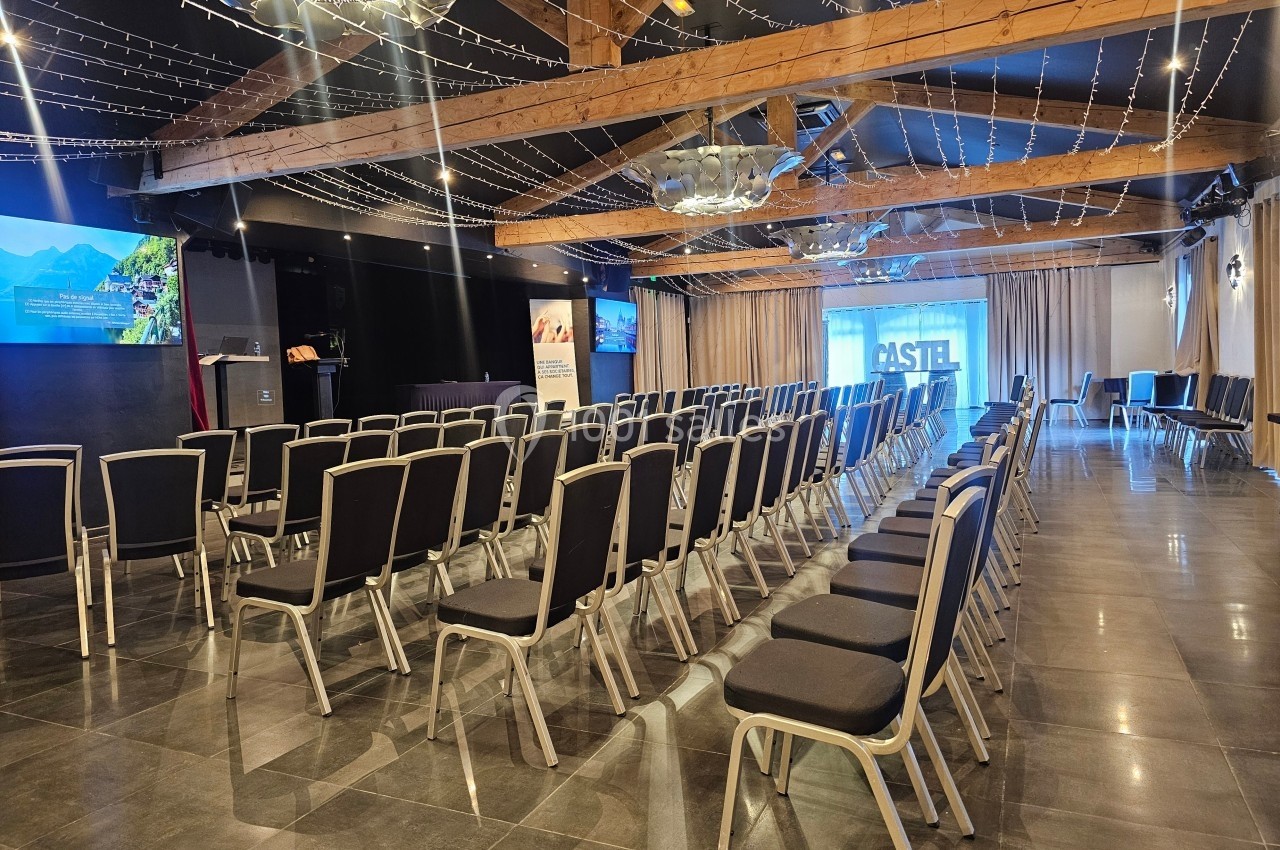 Salle de conférence avec rangées de chaises noires alignées, éclairage suspendu et écran de projection en fond.
