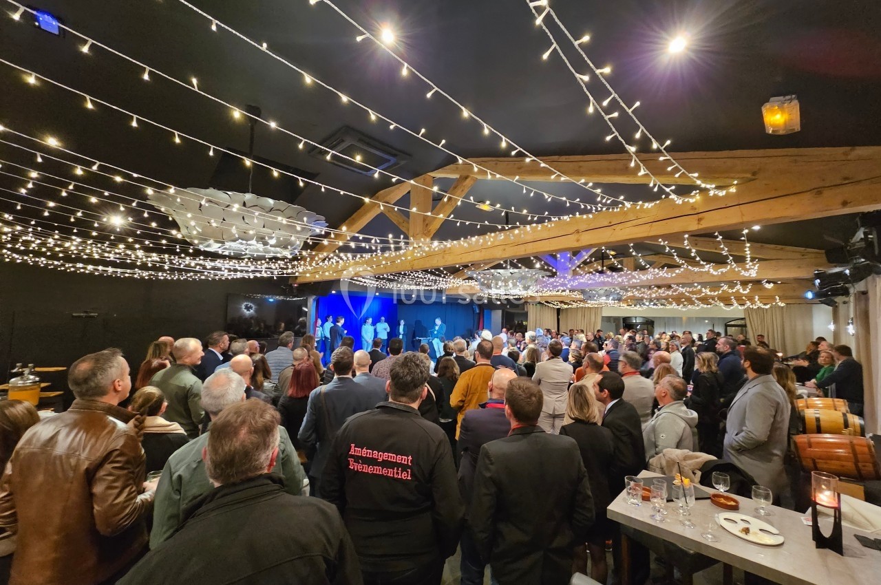 Salle remplie de personnes debout, éclairée par des guirlandes lumineuses, avec une scène en arrière-plan.