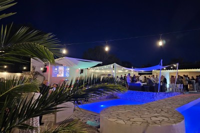 Un groupe de musiciens joue sur une petite scène extérieure sous un ciel bleu, entouré de décorations en bois.