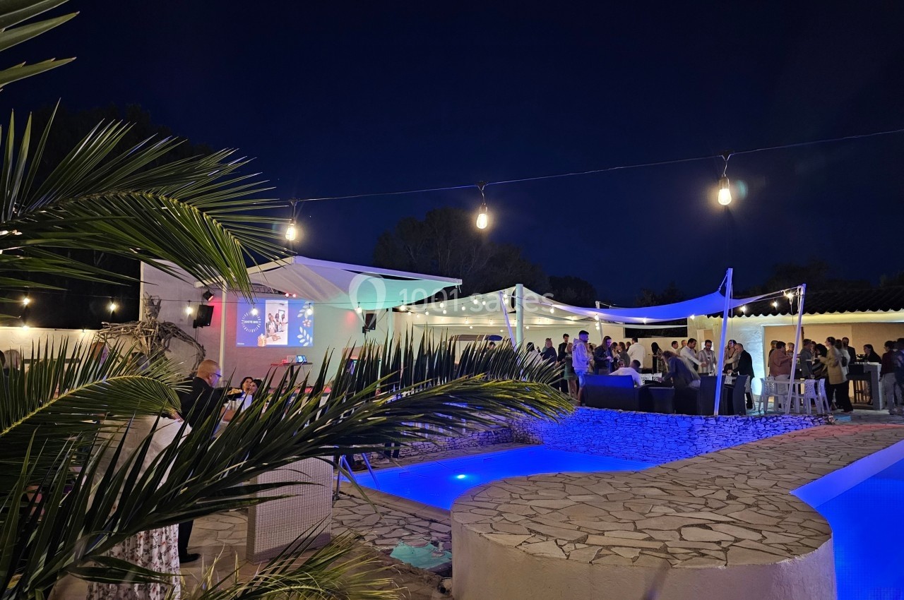Espace extérieur éclairé de nuit avec piscine, palmiers, et invités rassemblés sous des voiles blanches.