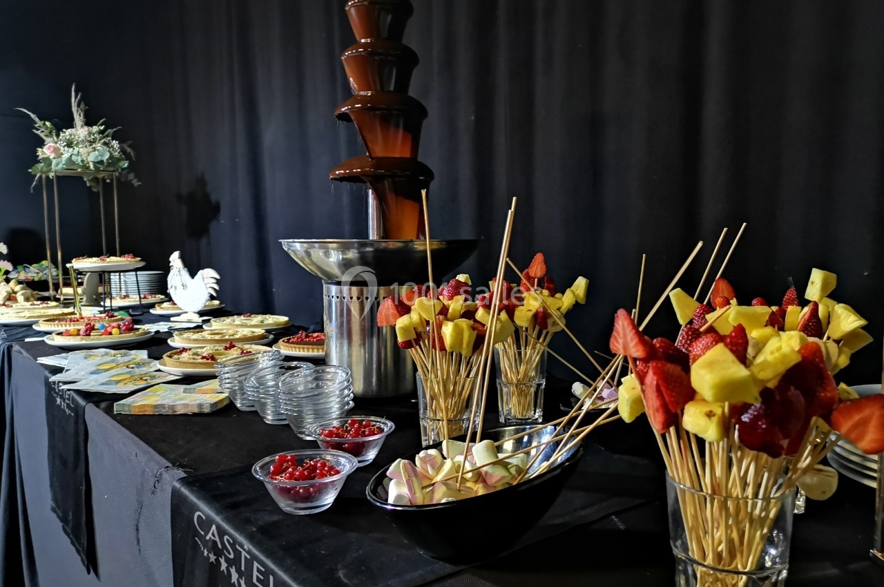 Fontaine de chocolat entourée de brochettes de fruits, desserts variés et décorations sur une table noire.