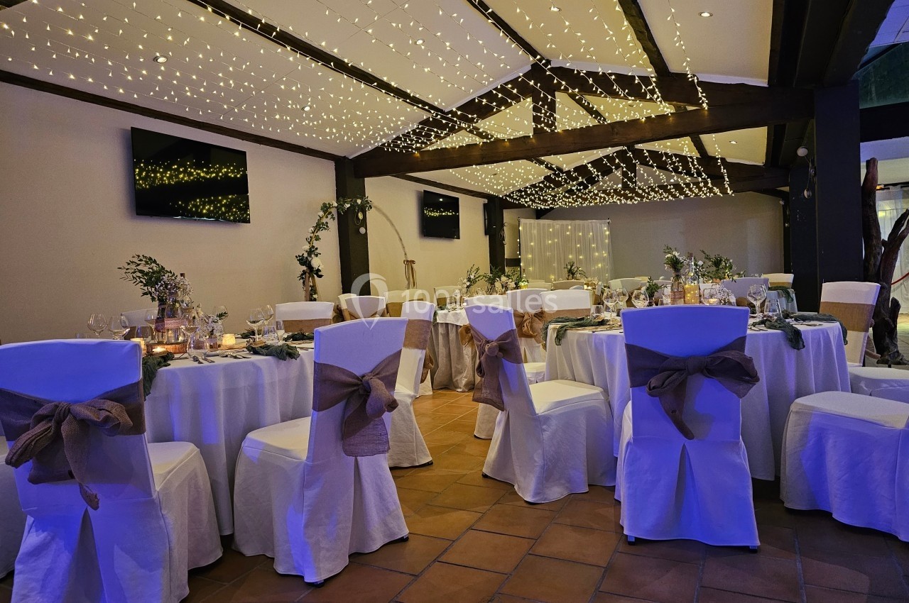 Salle de réception décorée avec des guirlandes lumineuses, tables rondes dressées et chaises ornées de nœuds en tissu.