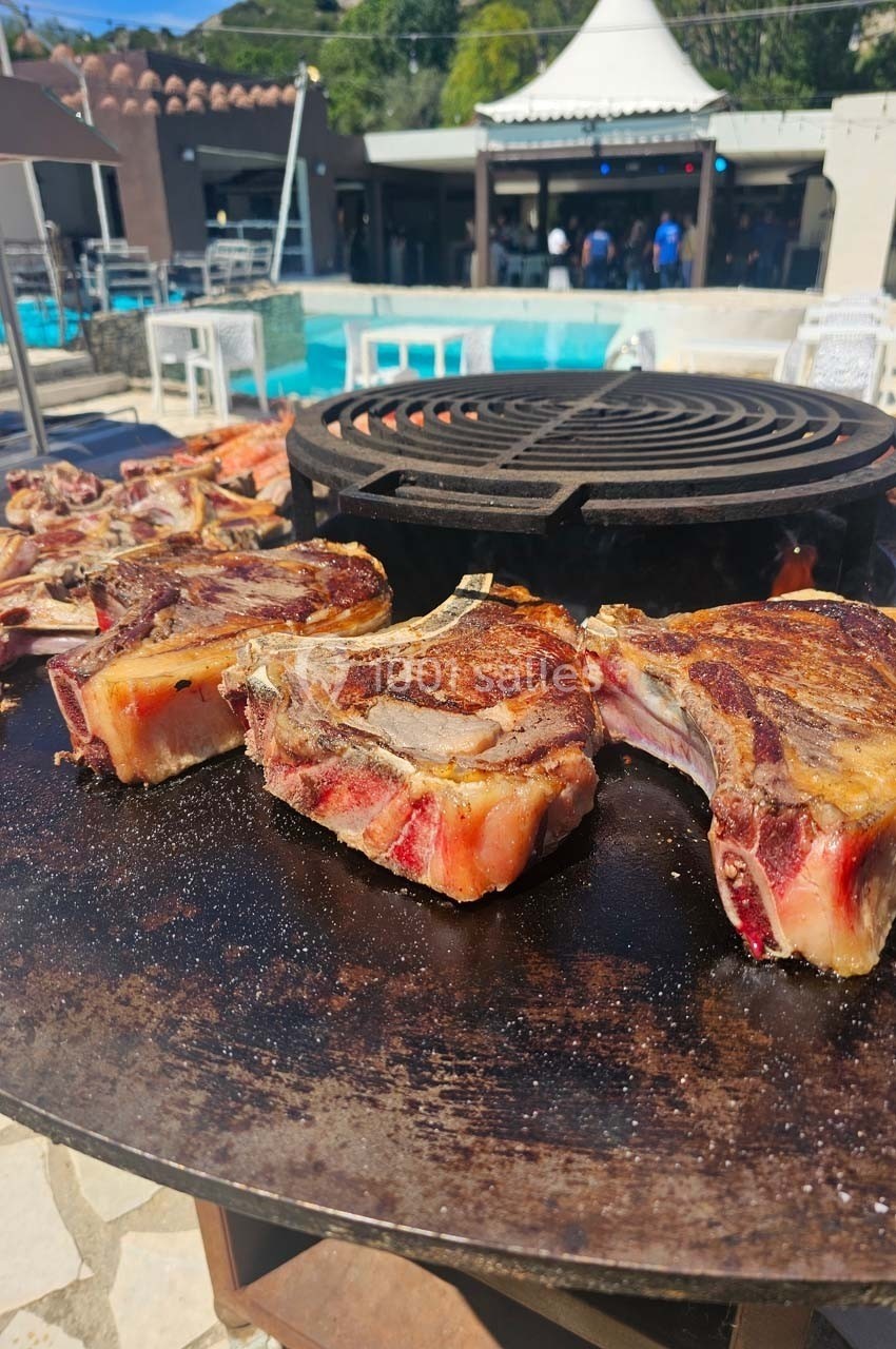 Côtes de bœuf grillant sur une plancha circulaire en extérieur, avec une piscine et des tables en arrière-plan.
