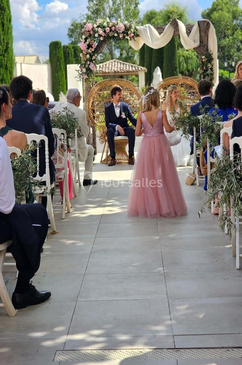 Une cérémonie de mariage en extérieur avec des invités assis, une fillette en robe rose marchant vers les mariés.