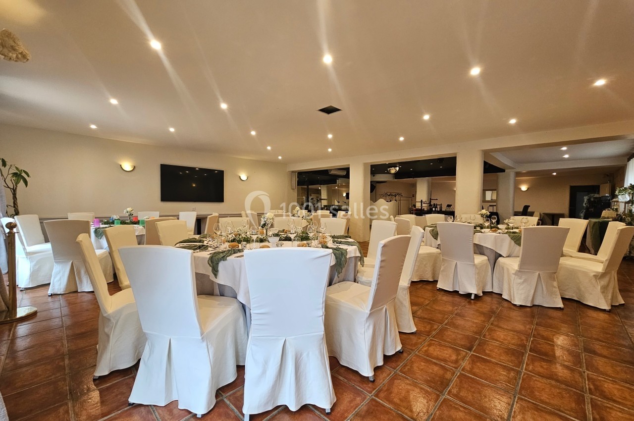 Salle de réception lumineuse avec des tables rondes dressées, entourées de chaises blanches et un sol en carrelage.