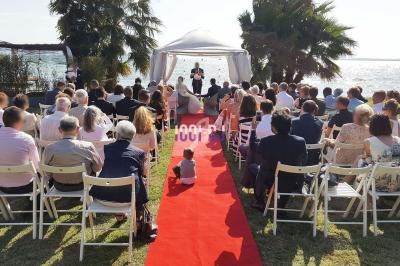 Cérémonie de mariage en plein air face à la mer, avec invités assis sur des chaises blanches et une allée rouge.