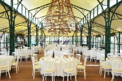 Salle de réception lumineuse avec tables rondes dressées, chaises blanches et grand lustre central décoratif.