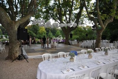 Scène extérieure avec piste de danse sous des arbres, décorée de lanternes blanches, entourée de tables dressées.
