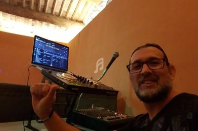 Un homme souriant pose devant une table de mixage, un ordinateur et un microphone dans une pièce au plafond en bois.