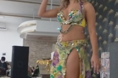 Femme en tenue colorée de danse orientale, souriante, debout dans une salle avec un décor moderne.