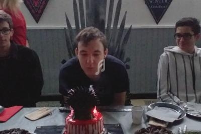 Un jeune homme souffle une bougie sur un gâteau décoré, entouré de deux personnes assises à une table.