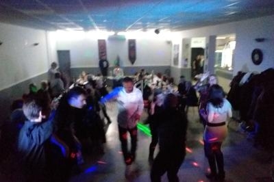 Personnes dansant dans une salle éclairée par des lumières colorées, avec des tables occupées en arrière-plan.