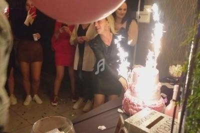Une femme souriante coupe un gâteau d'anniversaire décoré de bougies étincelantes, entourée de personnes.
