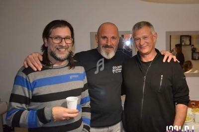 Trois hommes souriants posent ensemble dans une pièce éclairée, avec une table et des objets en arrière-plan.