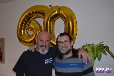 Deux hommes souriants posent devant des ballons dorés formant le chiffre 60, dans un intérieur décoré.