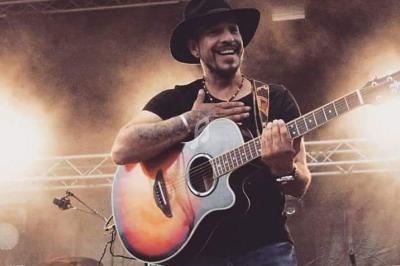 Un musicien souriant joue de la guitare acoustique sur une scène en plein air, entouré de fumée légère.