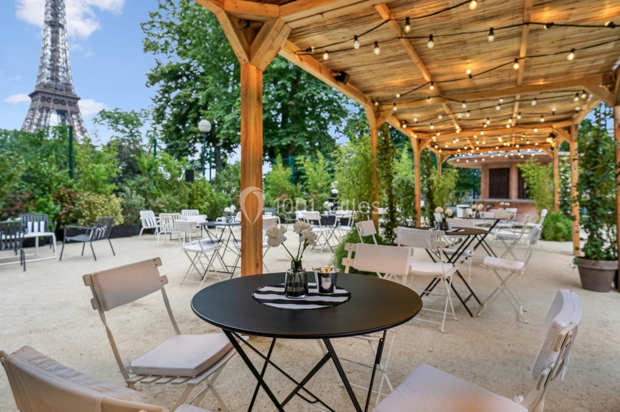 Terrasse extérieure avec tables et chaises sous une pergola éclairée, vue sur un jardin et la tour Eiffel en arrière-plan.