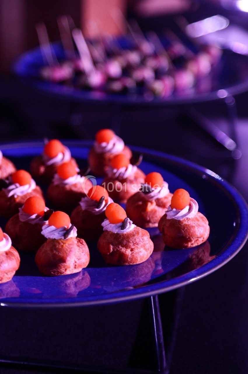 Plateau de petits choux garnis de crème et décorés de billes orange, avec des amuse-bouches flous en arrière-plan.