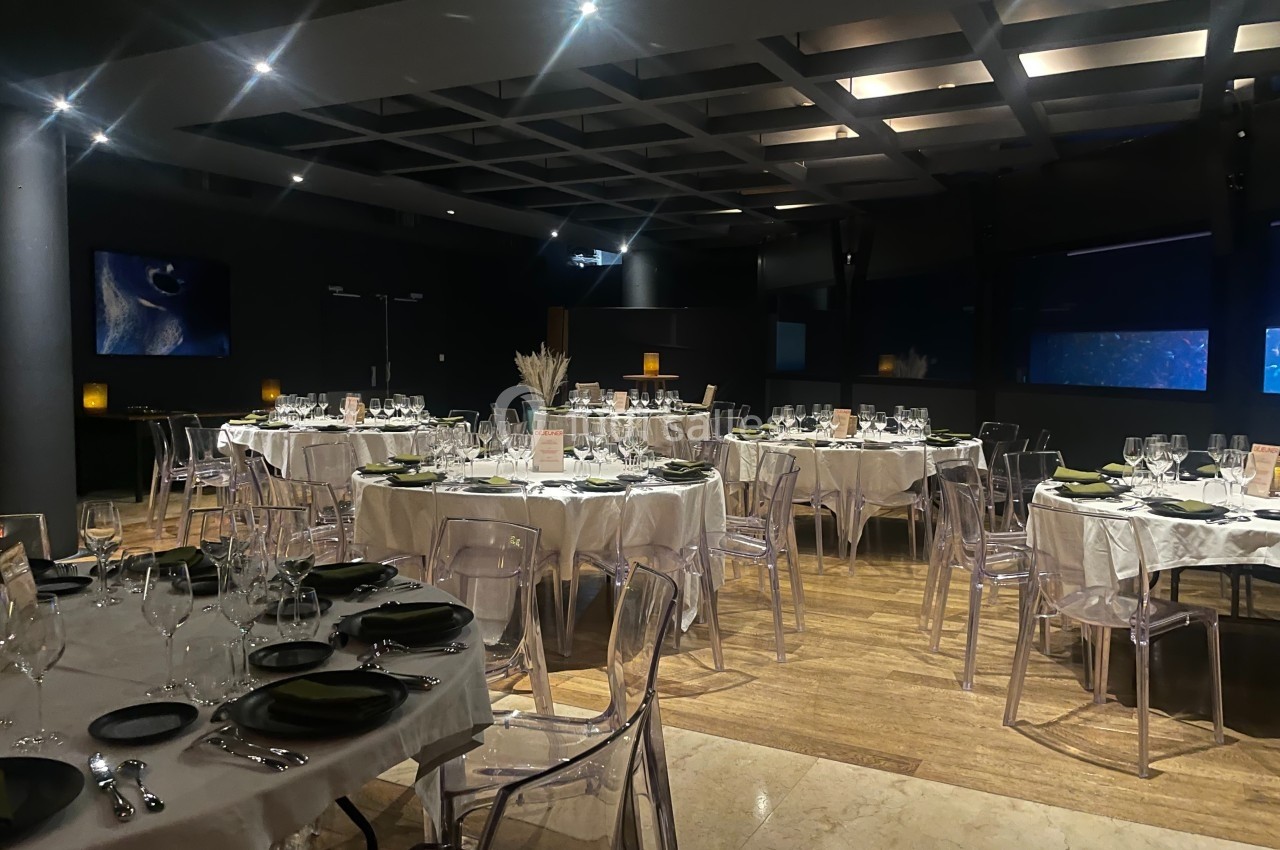 Salle de réception élégante avec tables rondes dressées, chaises transparentes et éclairage tamisé.