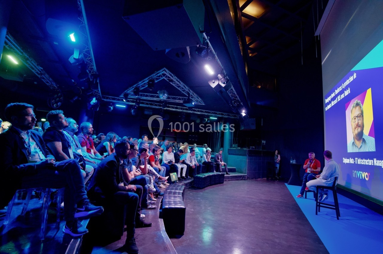 Un groupe de personnes assiste à une présentation dans une salle sombre avec un écran projetant un intervenant.