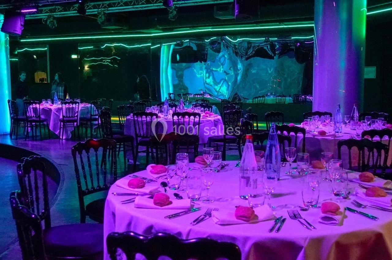 Tables rondes dressées avec nappes blanches, couverts et verres, dans une salle éclairée par des lumières colorées.