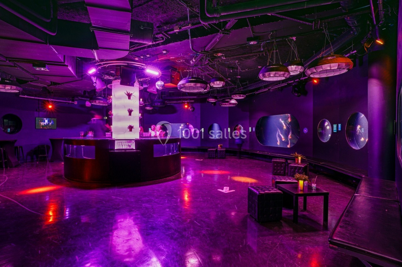 Salle sombre avec éclairage violet, bar central lumineux, tables basses et écrans ronds diffusant des images.